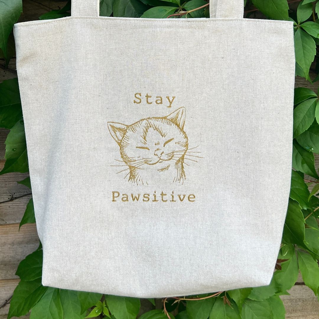 Tote bag with a cat illustration and text 'Stay Pawsitive' on a leafy background