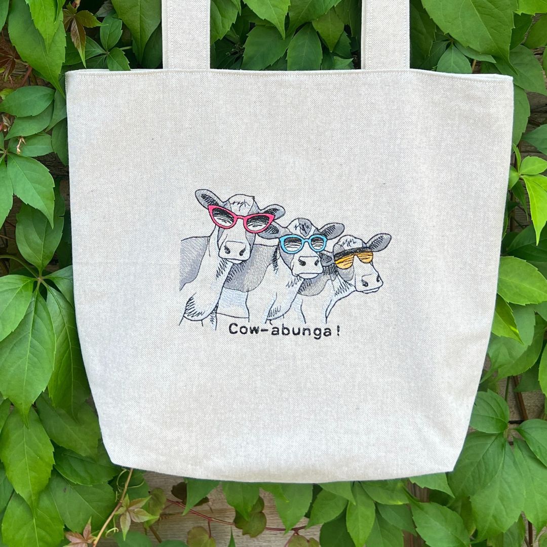 Tote bag with cow design and 'Cow-abunga!' text against a green leafy background