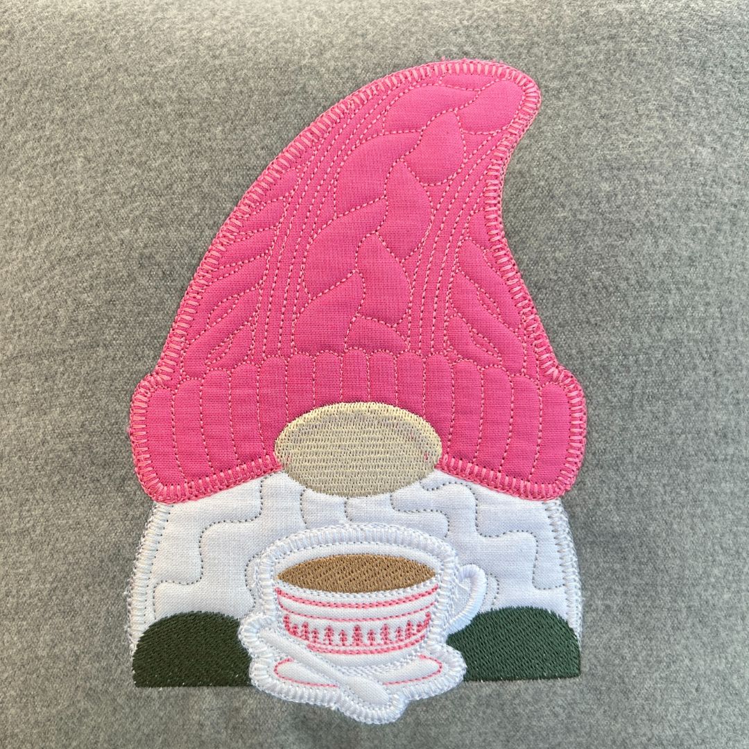 Embroidered patch of a gnome with a pink hat holding a cup on a gray fabric background