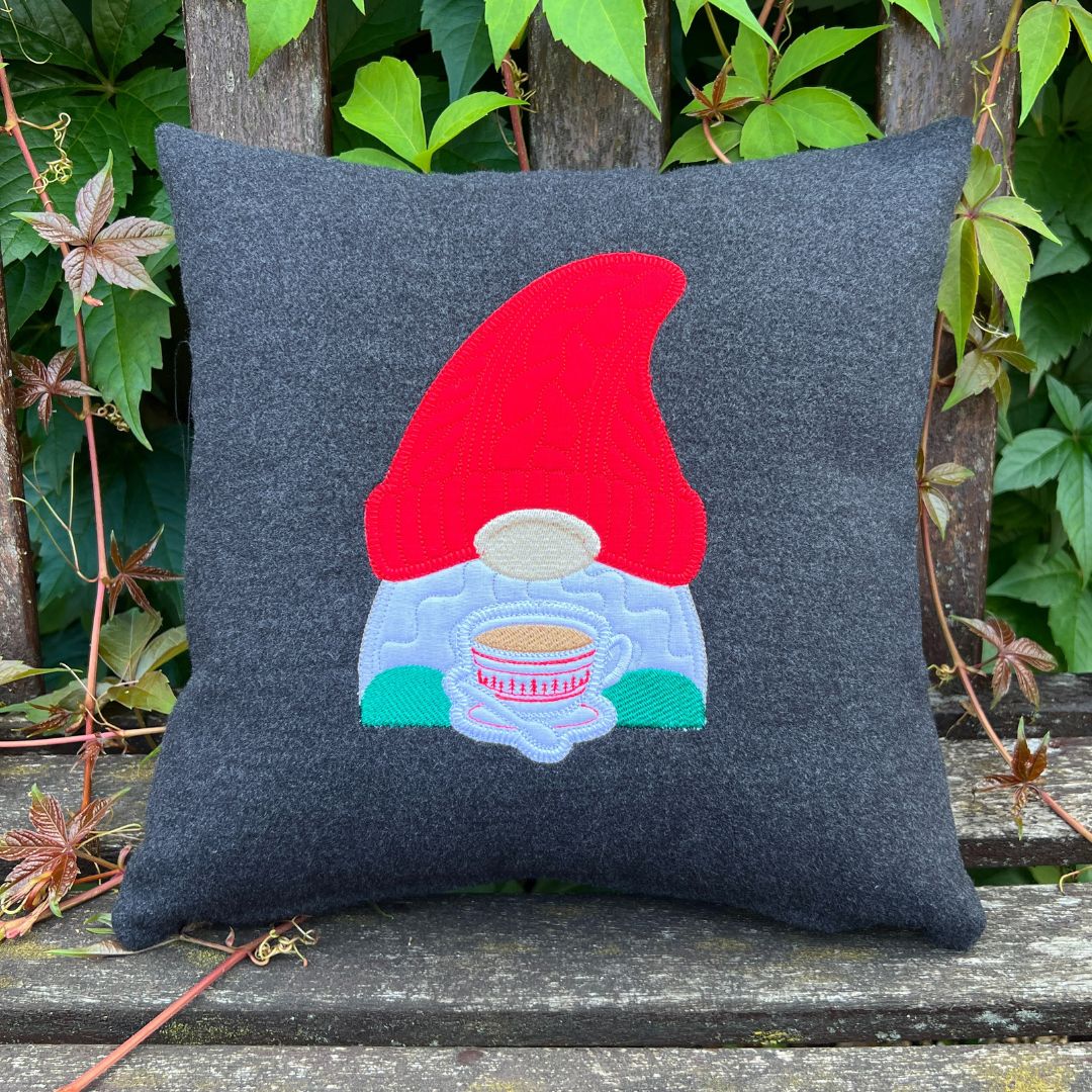 Gray pillow with a gnome design holding a cup on a wooden surface with greenery.
