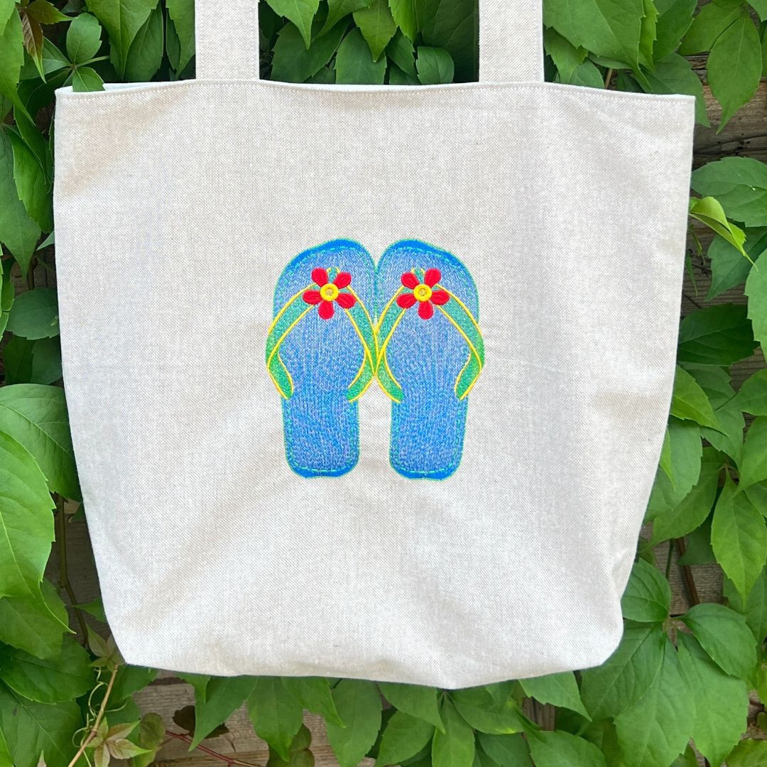 Beige tote bag with blue flip-flop design featuring flowers against a green leafy background
