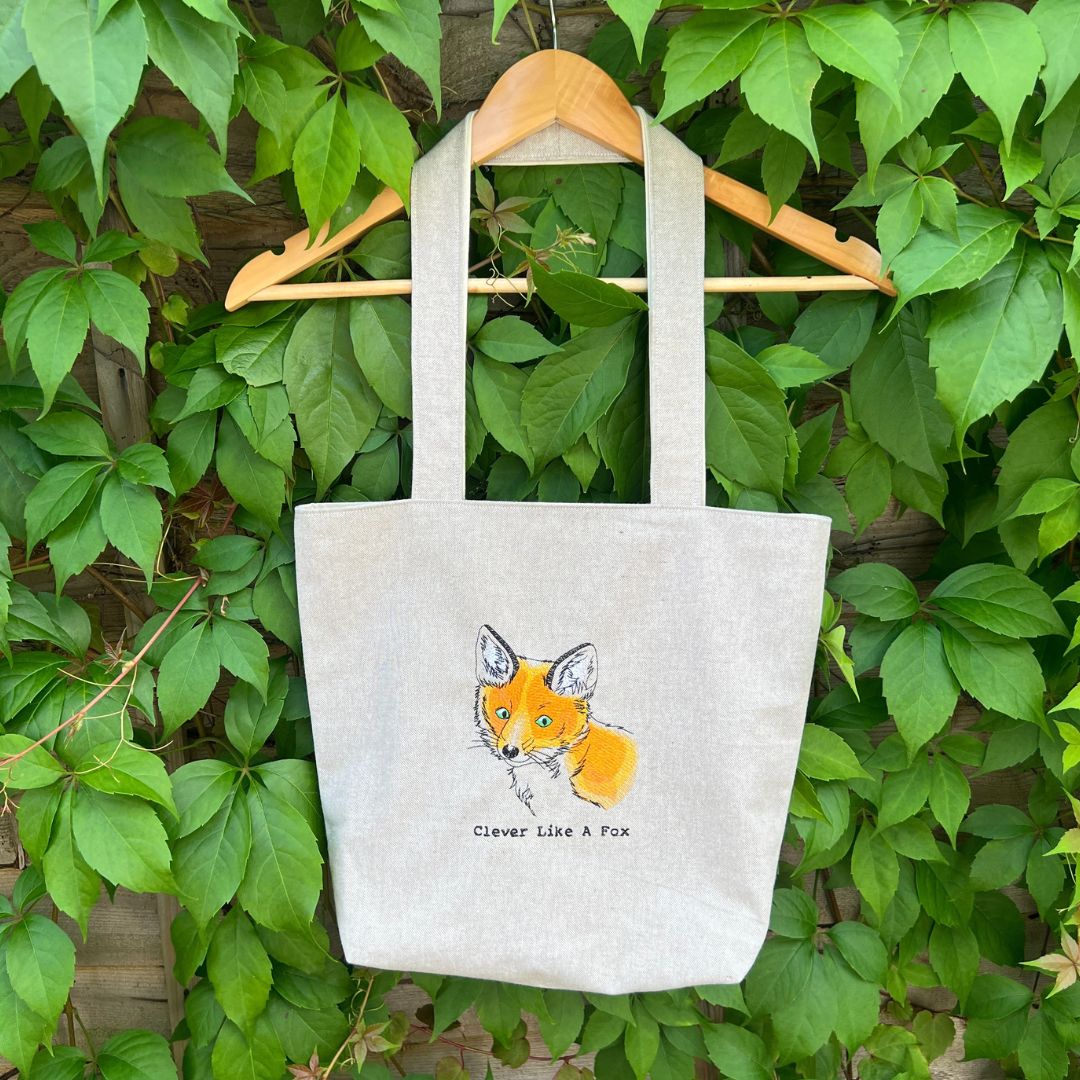 Tote bag with fox design and text 'Clever Like A Fox' hanging on a wooden hanger against a green leafy background.