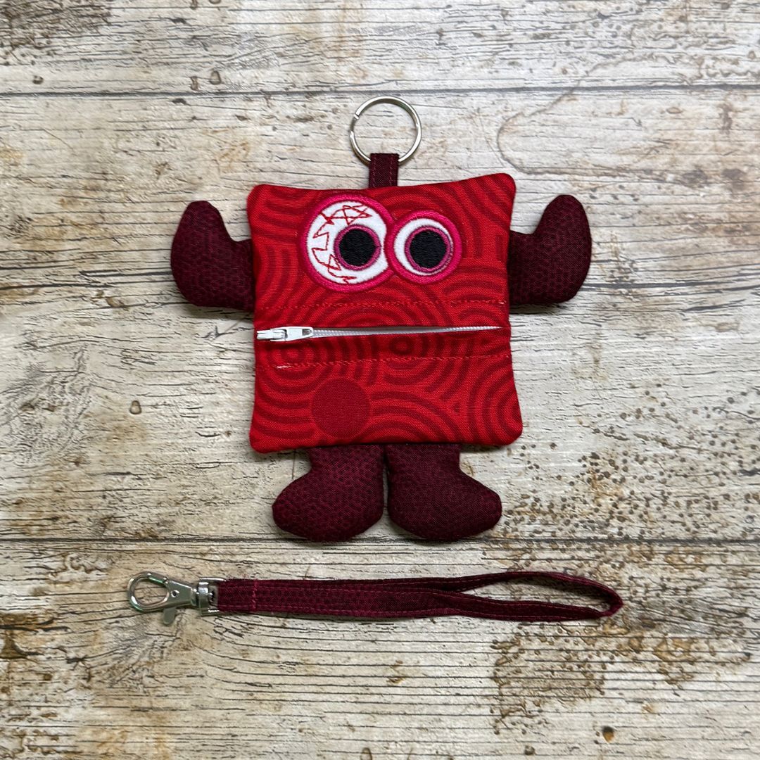 Red Money monster called Swirlie Stash with a keychain and a zipper for a mouth, plus a wrist strap on a wooden surface