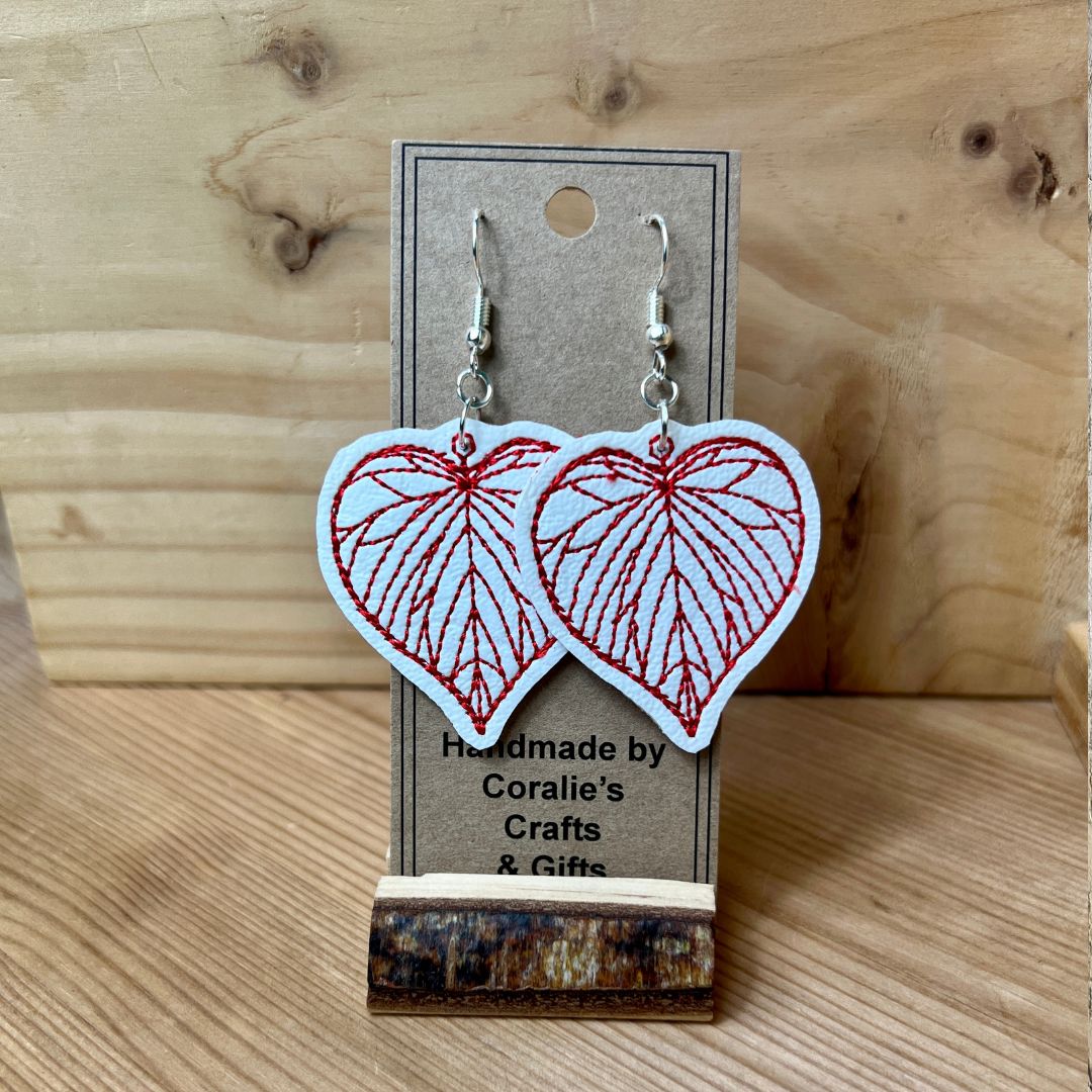 A pair of heart-shaped dangle earrings with red embroidered leaf patterns on a white background, hanging from silver fishhook wires on a brown kraft display card. The card is placed on a rustic wooden block over a light wood grain surface.