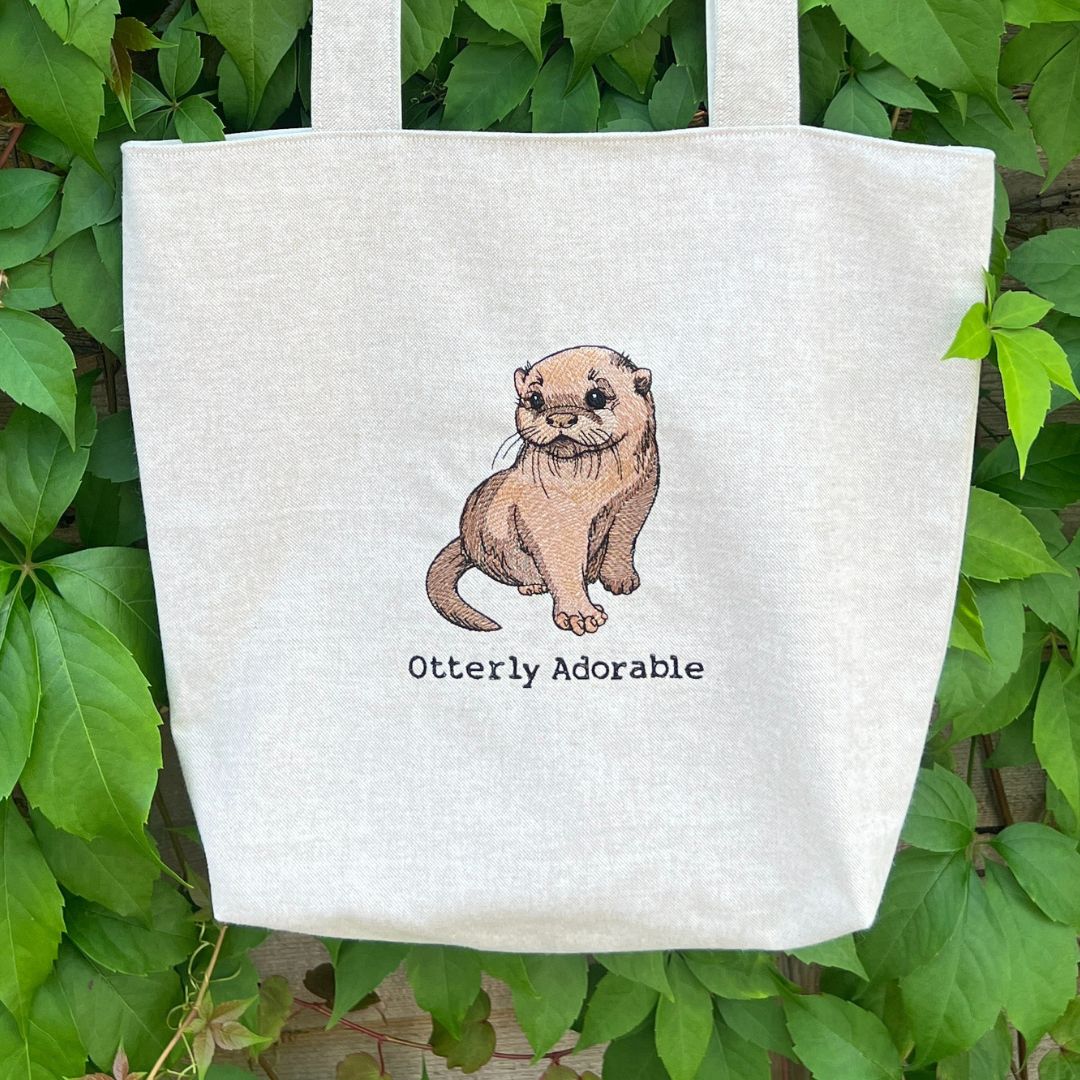 White tote bag with an otter illustration and 'Otterly Adorable' text, against a green leafy background