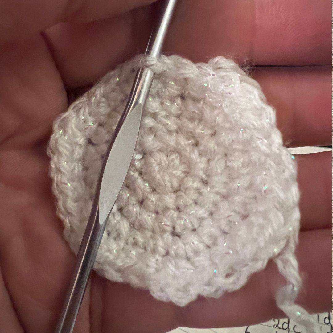A close-up of a hand holding a small crocheted round piece in white yarn with a silver crochet hook inserted.