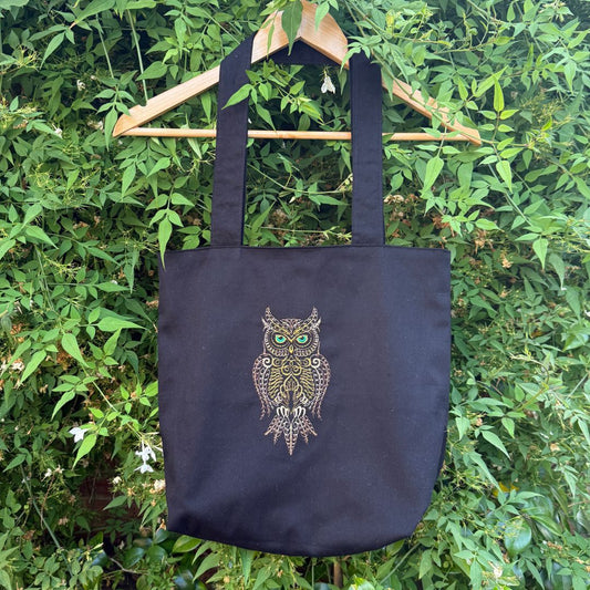 A black canvas tote bag with a detailed embroidered brown owl graphic hanging from a wooden hanger against a background of green foliage.