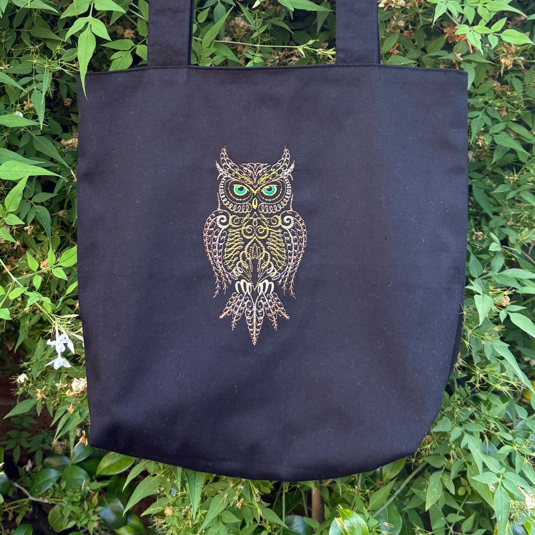 A black canvas tote bag with a central embroidered brown owl design featuring green eyes and gold and brown patterns, set against a background of green foliage.