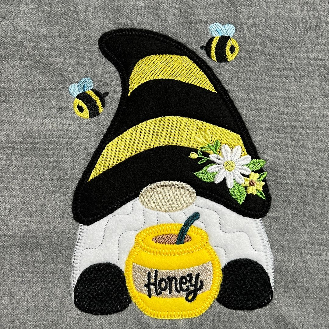 Embroidered patch of a gnome with a honey pot and bees on a gray fabric background