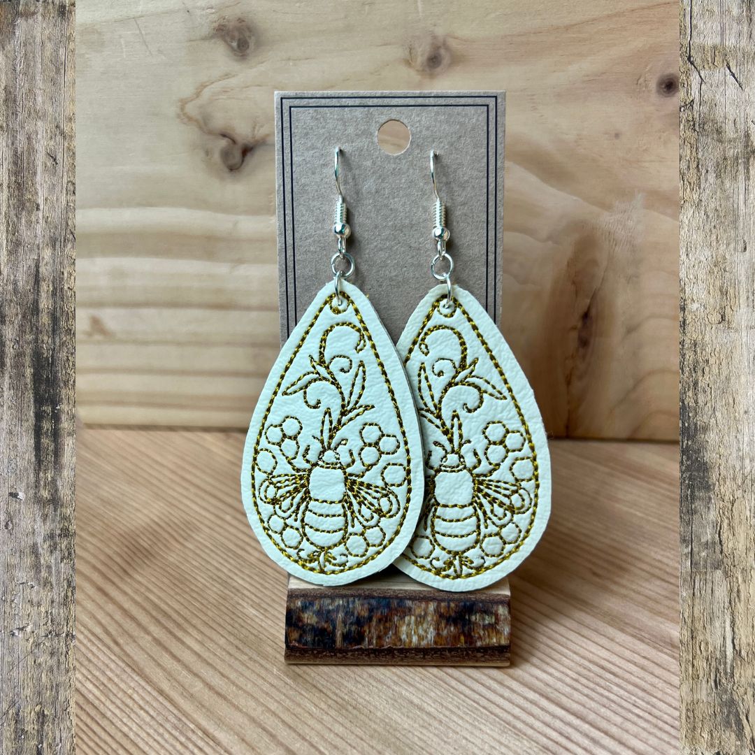 A pair of dangling teardrop-shaped earrings with a pale mint green background and olive green embroidered floral and bee designs, displayed on a kraft card stand against a rustic wood background.