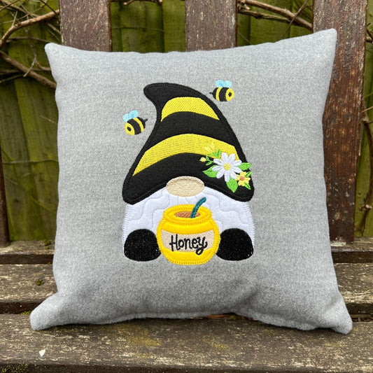 Gray pillow with a bee-themed gnome design featuring a black hat, yellow body, and a honey jar on a wooden surface.