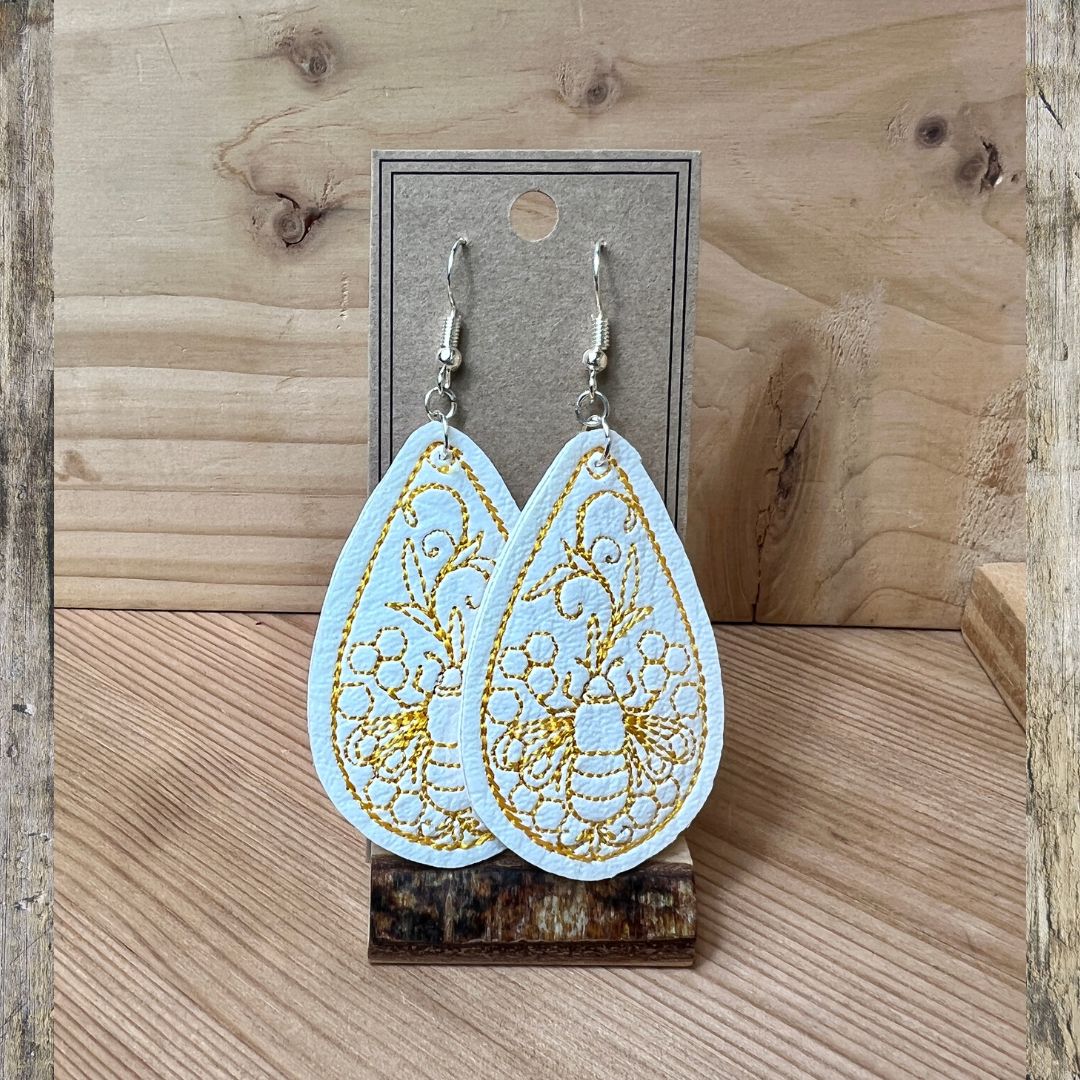 A pair of light blue teardrop-shaped dangle earrings with gold embroidered floral patterns, displayed on a gray card stand against a rustic wooden background.