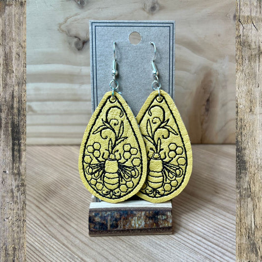 A pair of yellow teardrop-shaped dangle earrings with black embroidered floral and bee designs, displayed on a kraft card stand against a rustic wood background.