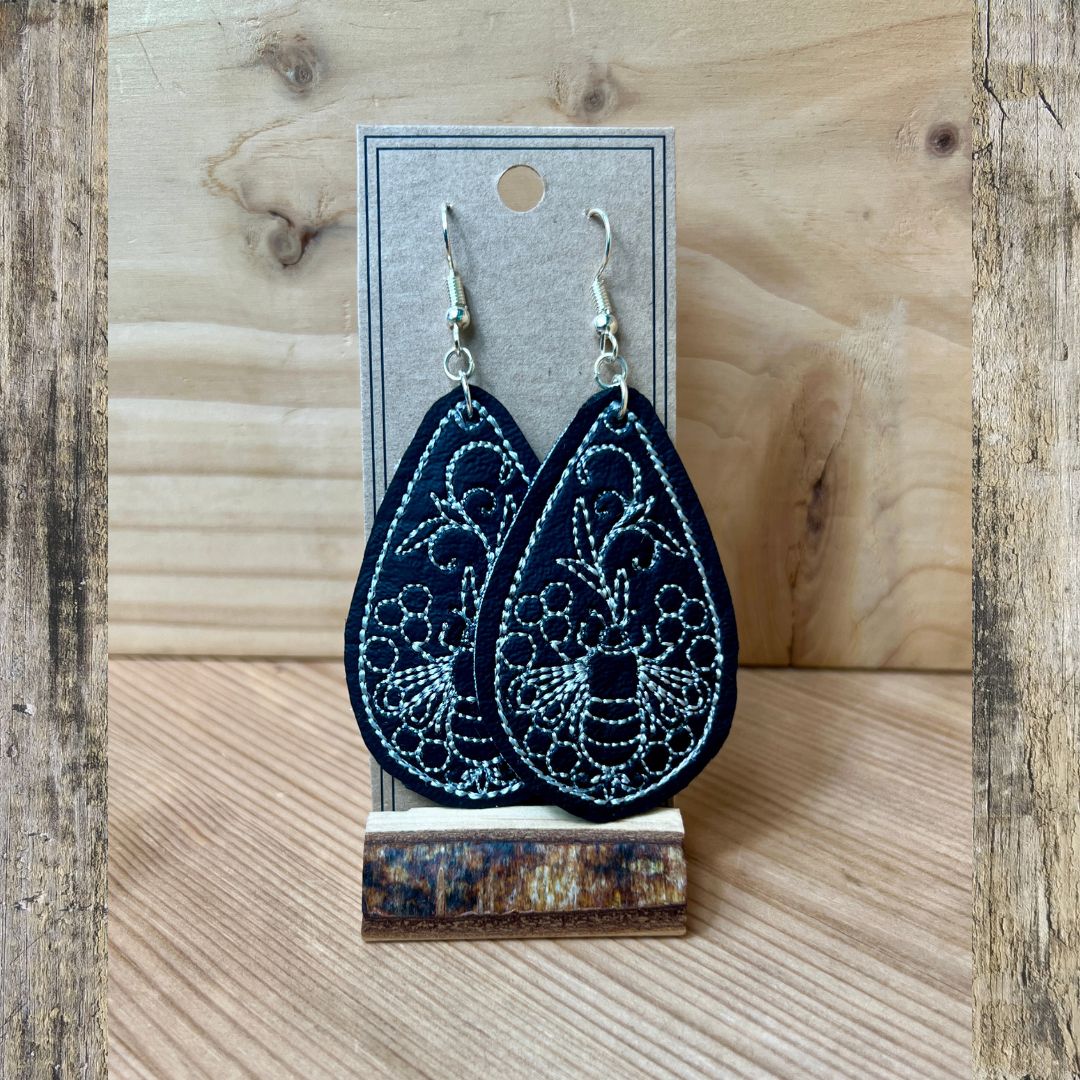 A pair of dark navy black teardrop-shaped fabric earrings with white embroidered floral patterns and silver fishhook ear wires, displayed on a kraft card and rustic wood block stand against a light wood background.