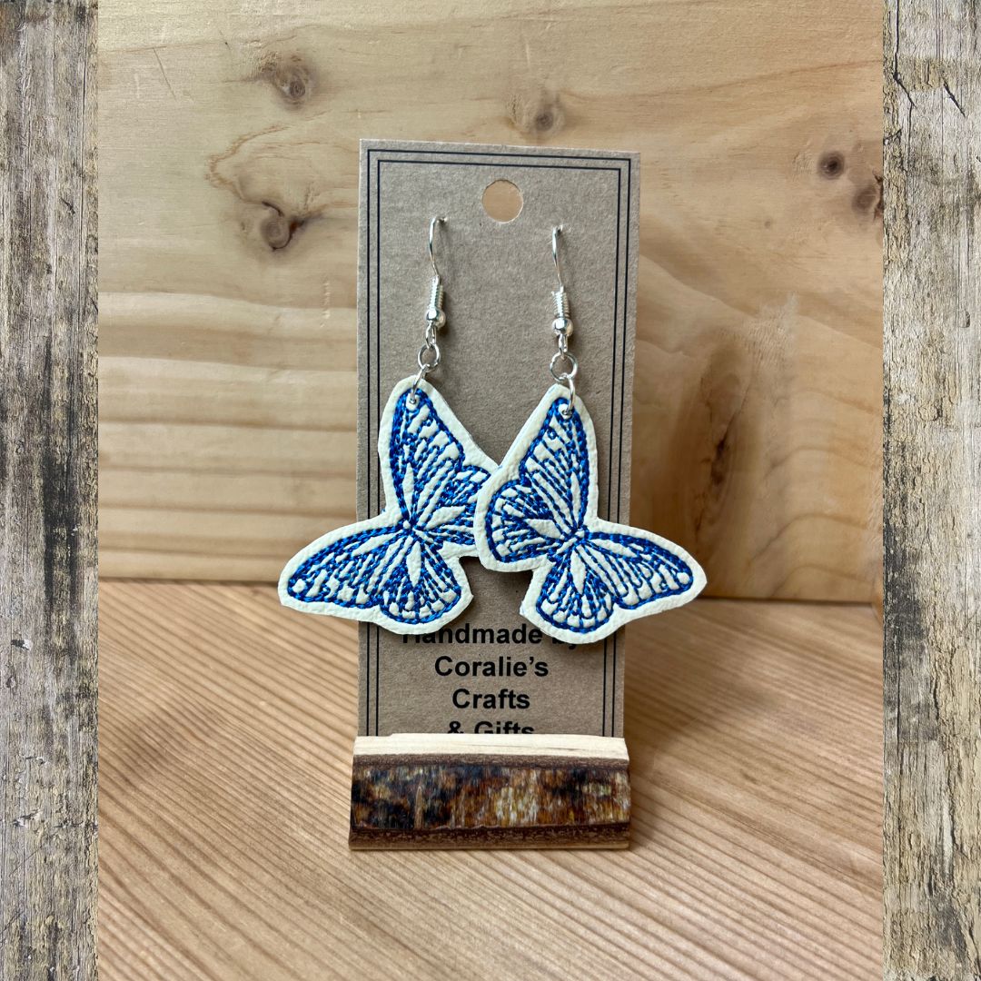 A pair of butterfly-shaped dangle earrings with blue embroidered details on a white fabric base, hanging from silver hooks on a brown kraft display card. The card is propped on a small wooden stand against a light wood background.