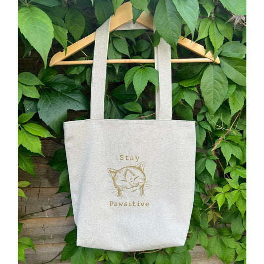White tote bag with 'Stay Pawsitive' text and cat illustration hanging on a green leafy background