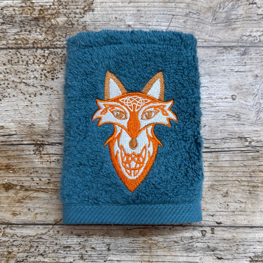 Blue towel with an embroidered fox design on a wooden surface. Embroidered at Coralie's Crafts & Gifts in Dorset.