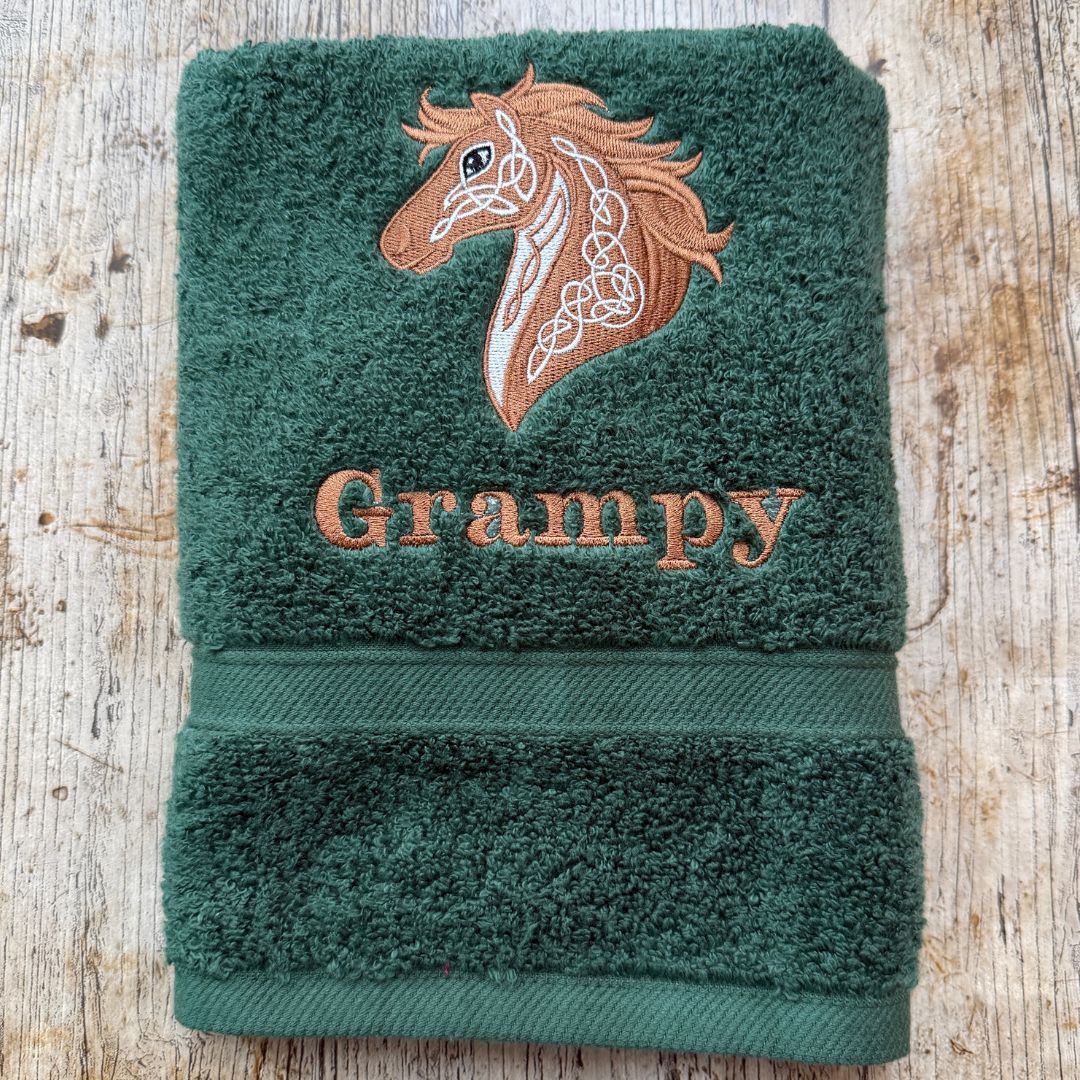 A folded Fir Green hand towel with an embroidered brown horse head and the name 'Grampy' in matching brown embroidery, displayed on a rustic light wood background.