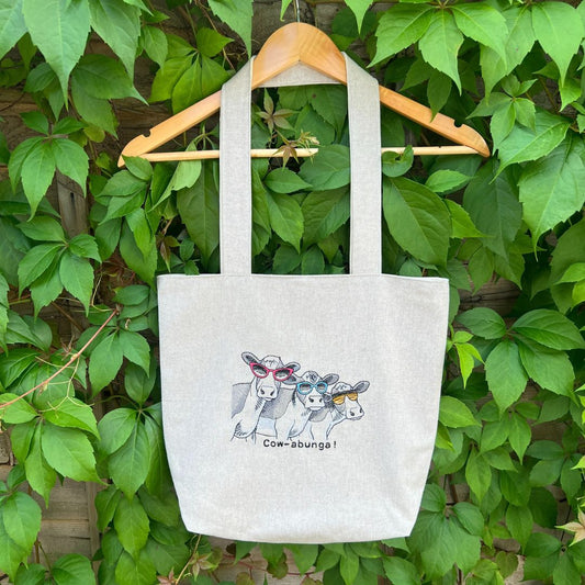 Tote bag with cow design hanging on a wooden hanger against a green leafy background