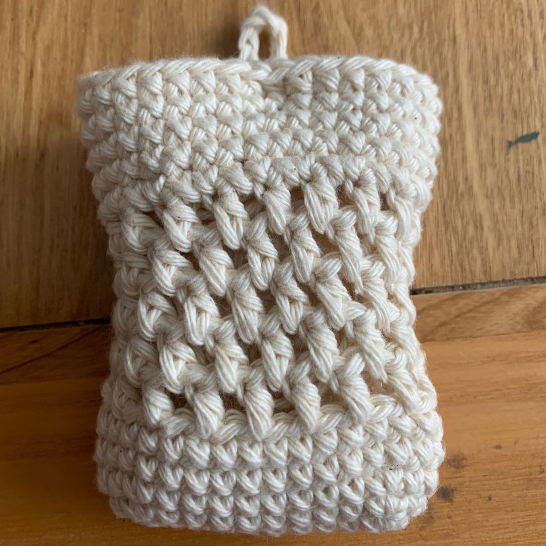 A handmade crocheted soap cozy with a textured crochet pattern and a small hanging loop at the top, resting on a wooden surface.