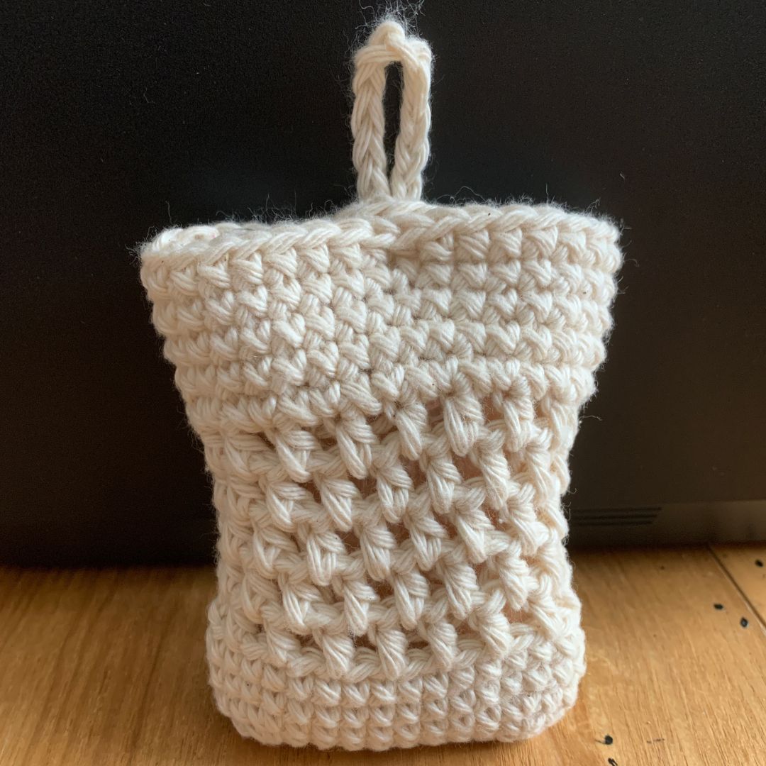 A cream-colored crocheted soap cozy bag with a hanging loop, standing upright on a wooden surface against a dark background.