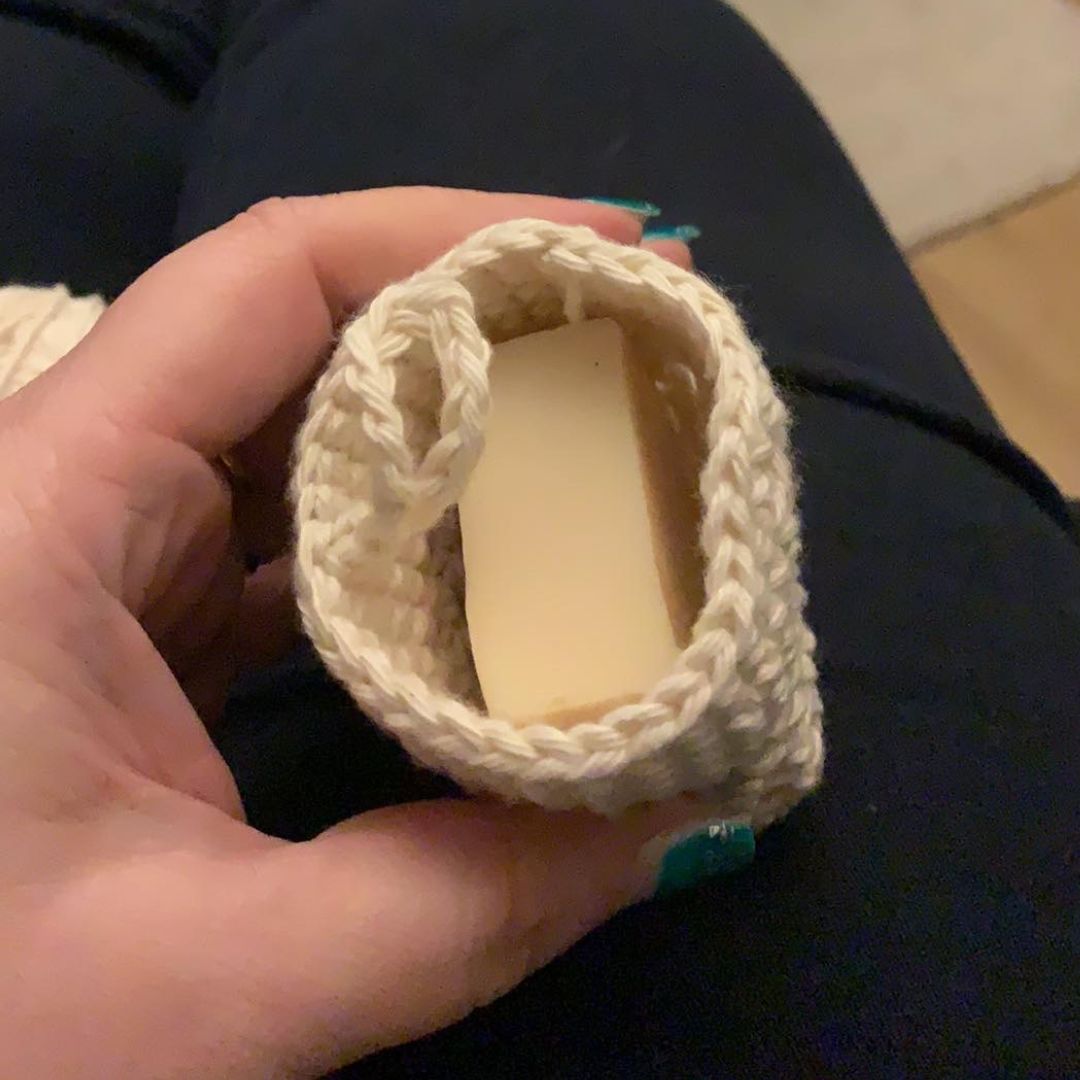 A hand with teal nail polish holds a small handmade cream-colored crocheted Soap Cozy containing a bar of soap, set against dark background