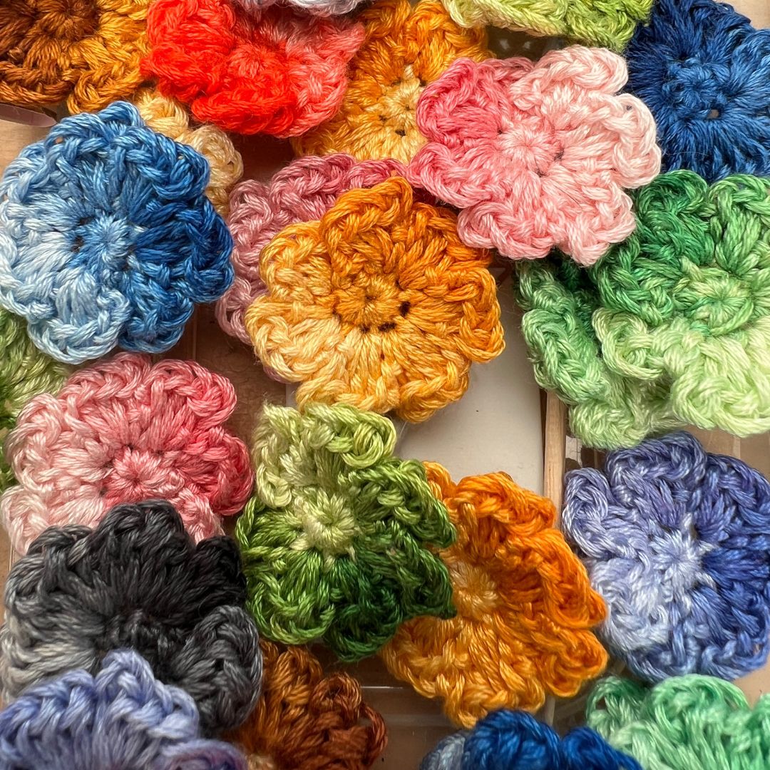 A close-up arrangement of colorful crocheted flower petals or small fabric flowers in many shades including blue, pink, orange, green, purple, brown, and gray, filling the frame.
