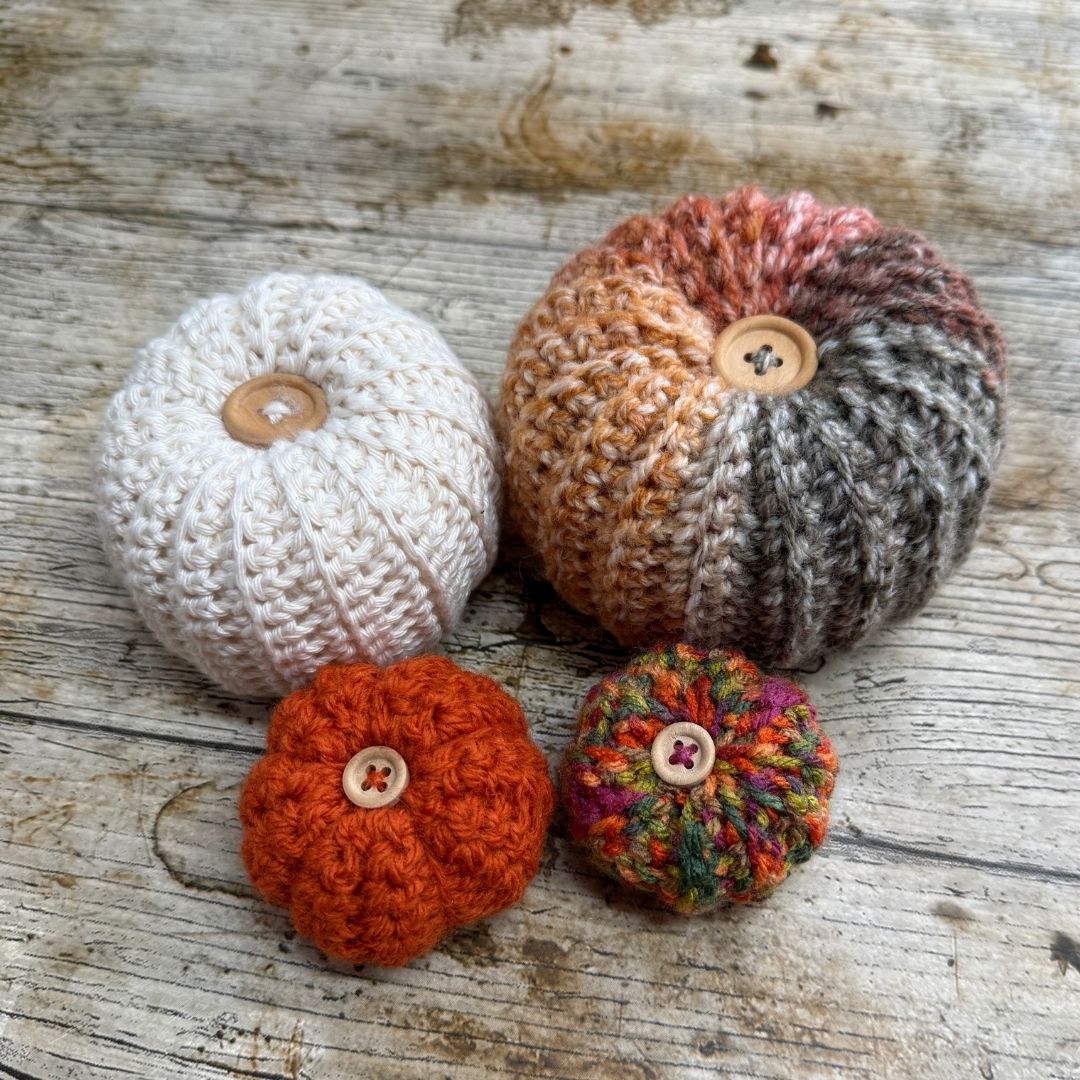 Four hand-crocheted mini pumpkins arranged on a rustic weathered wood surface. The pumpkins come in various colors including cream, orange, rust, and multicolor, each featuring a small wooden button on top.