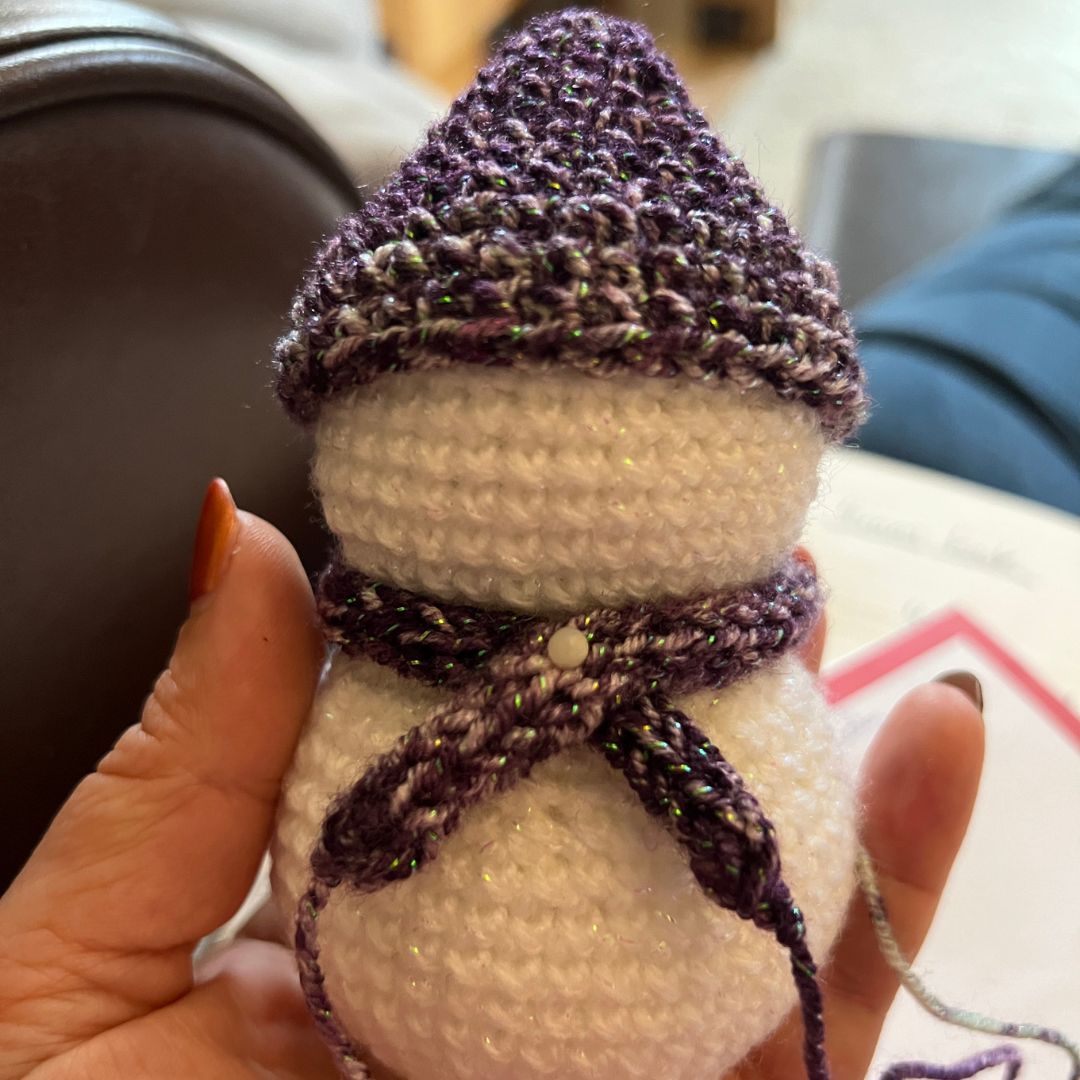 A handmade crocheted snowman toy held in a person's hand. The snowman has a white body with a pointed purple and white glittery hat and a matching purple and white glittery scarf.