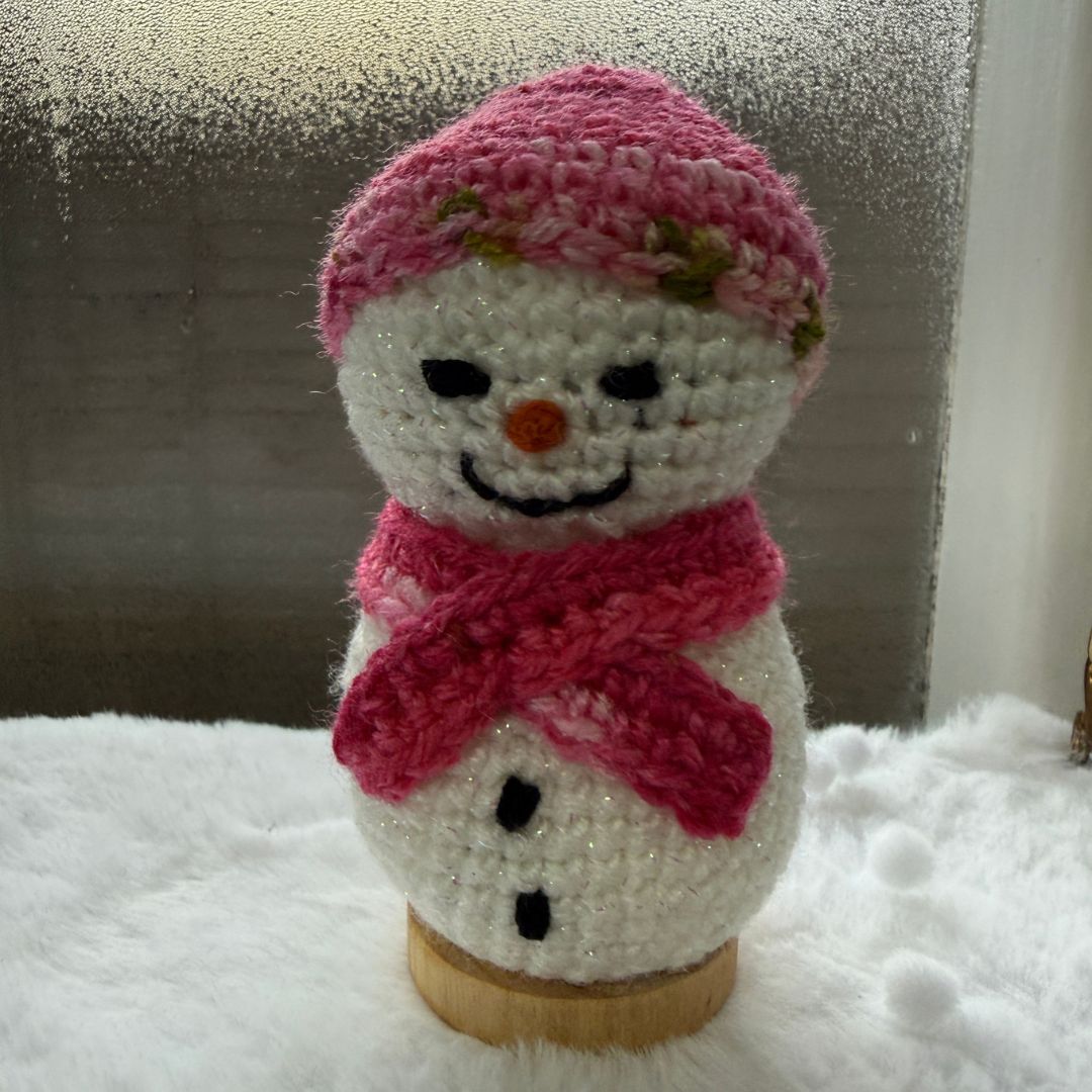 A crocheted snowman toy or ornament with a pink hat, pink scarf, black embroidered facial features, and an orange carrot nose, standing on a small wooden base against a frosted window and white fluffy surface.