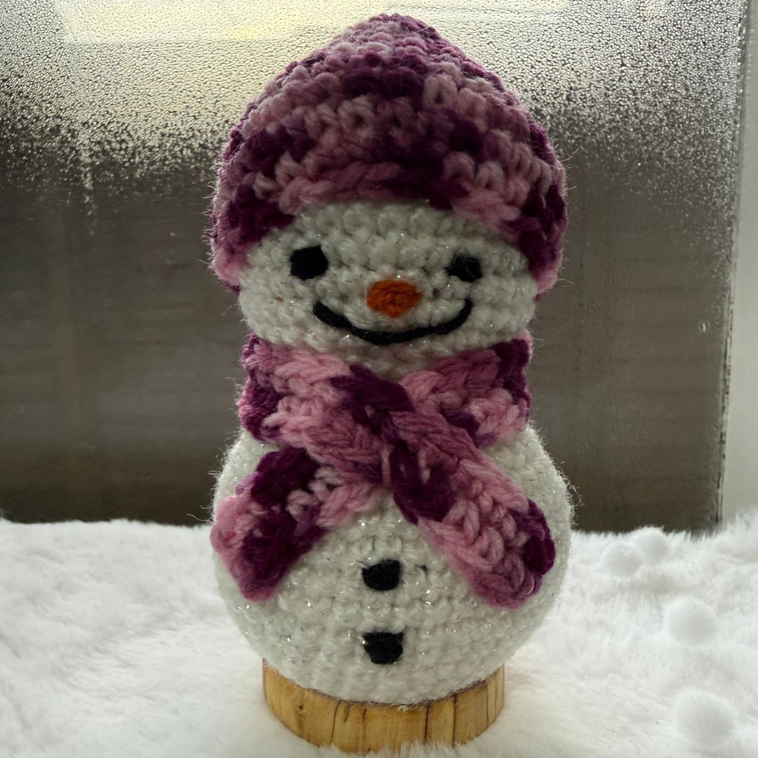 A crocheted snowman decoration with a white body, black facial features, an orange carrot nose, and a purple and pink knit hat and scarf, displayed on a small wooden base against a snowy window backdrop.
