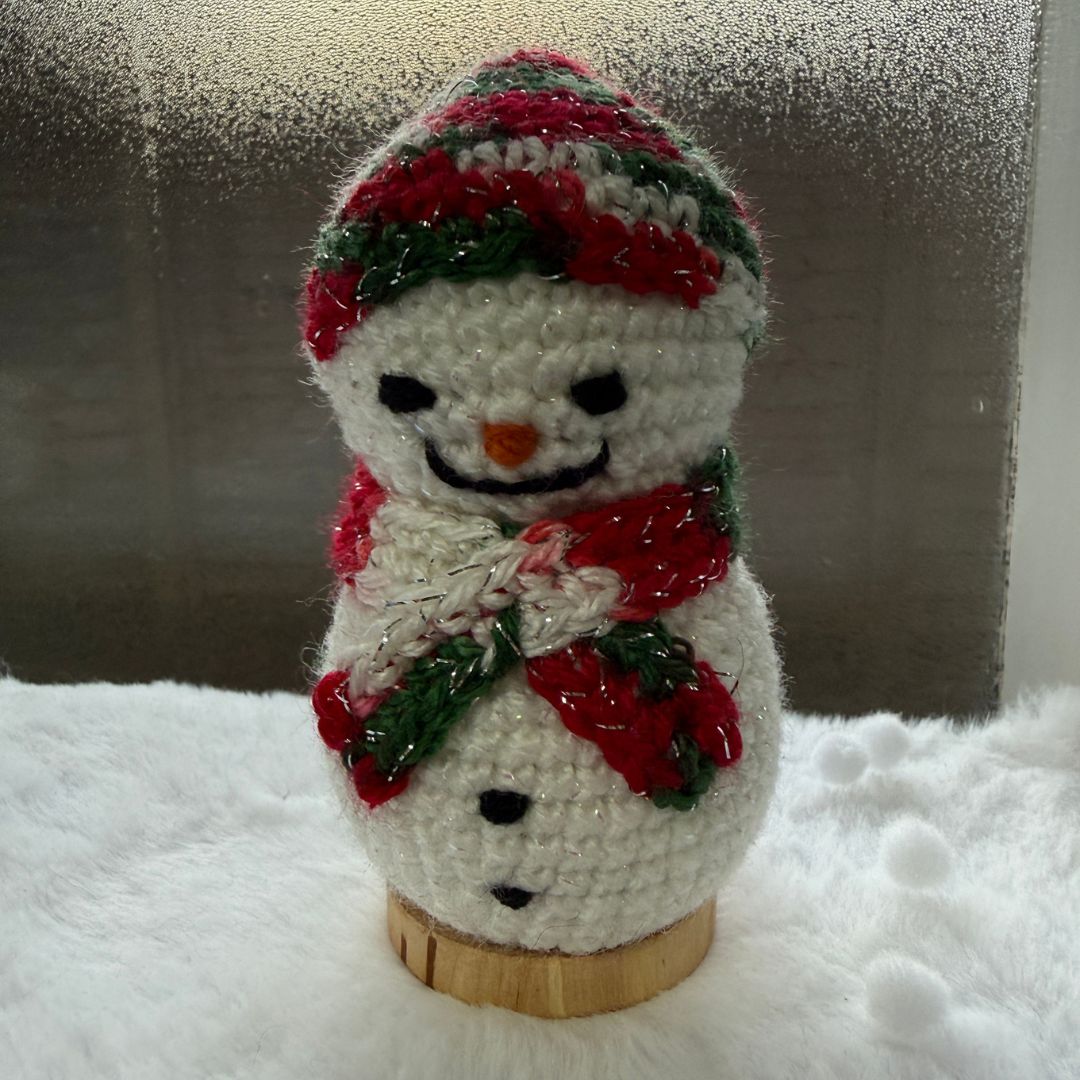 A handmade crocheted snowman ornament on a small wooden base, featuring a red, green, and white striped hat, a red and green scarf, and a black embroidered face, set against a snowy white backdrop and a textured gray background.