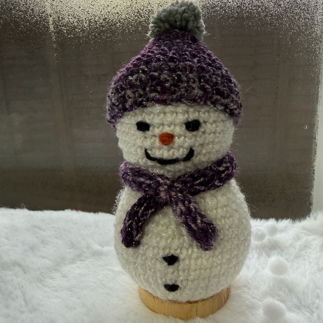 A handmade crocheted snowman figure wearing a purple knit hat with a pom-pom and a matching purple scarf, standing on a small wooden base against a snowy white surface and a gray textured background.