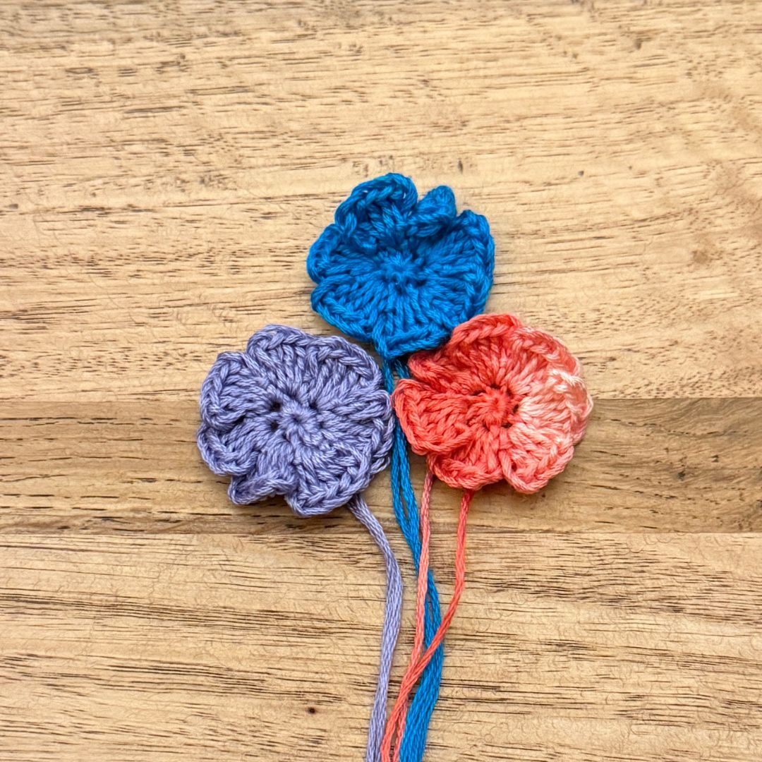 Three crocheted flowers in blue, purple, and pink on a wooden surface