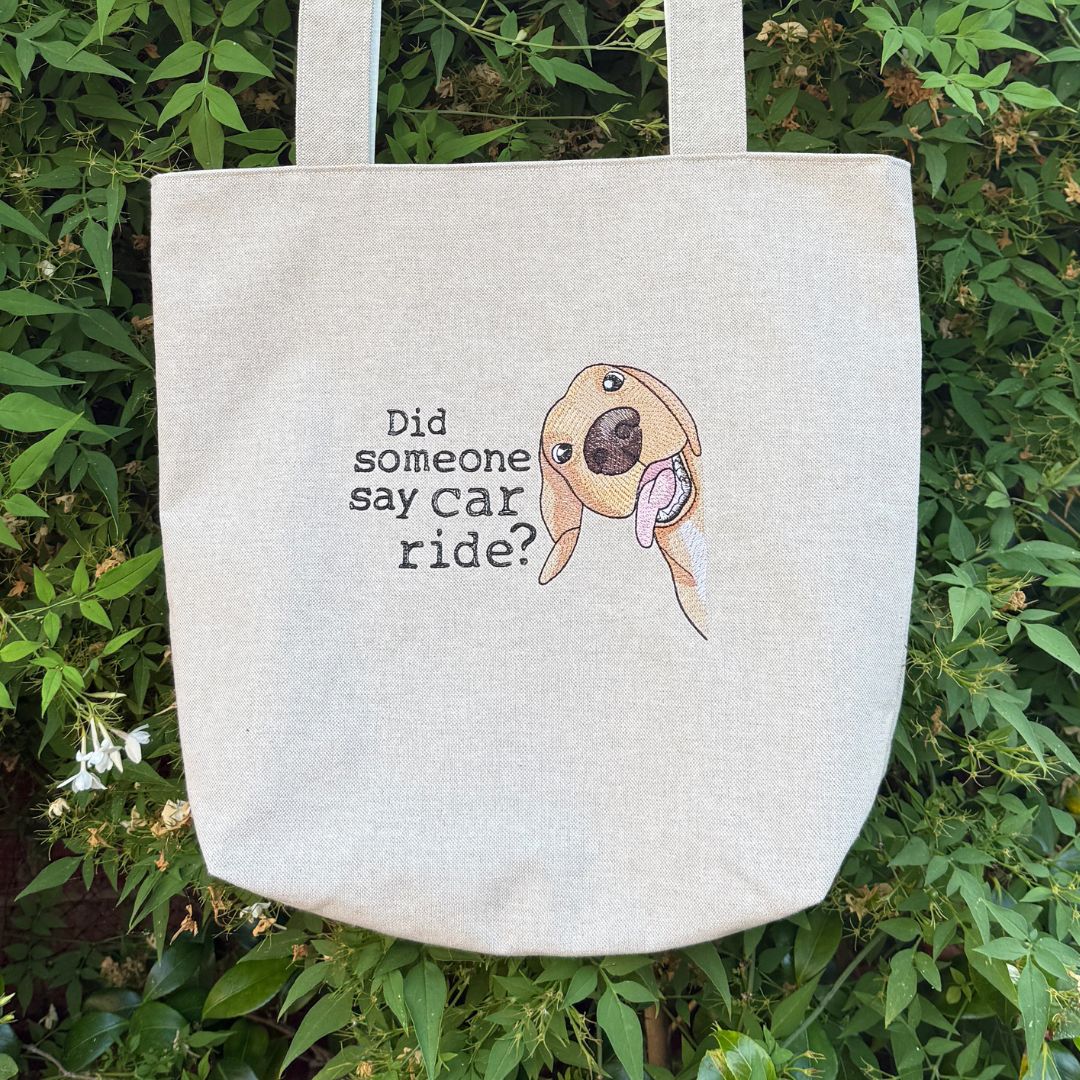 A natural canvas tote bag with a printed cartoon dog face and the text 'Did someone say car ride?' resting on green foliage.