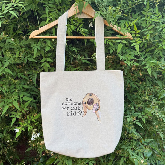 A natural canvas tote bag hanging from a wooden hanger against a background of green foliage. The bag features a printed graphic of a cartoon dog wearing sunglasses next to the text 'Did someone say car ride?'.
