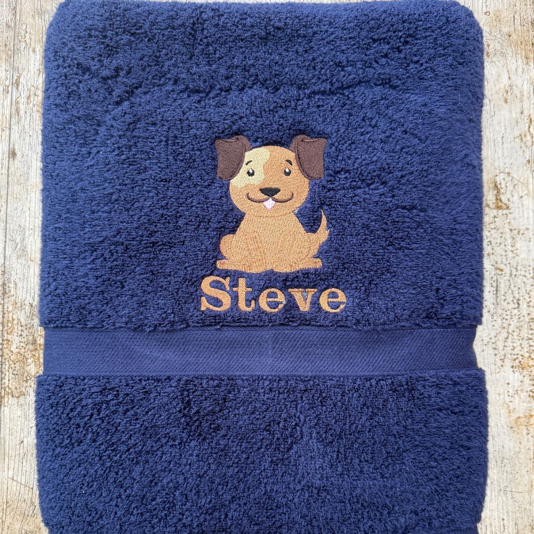 A folded navy blue Bath Sheet towel with an embroidered brown dog and the name 'Steve' in matching thread, laid flat on a light wood background.