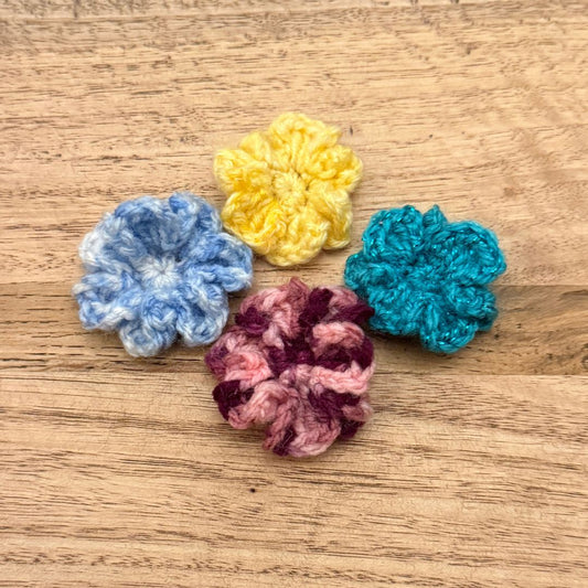 Four crocheted flowers in blue, yellow, pink, and teal on a wooden surface.