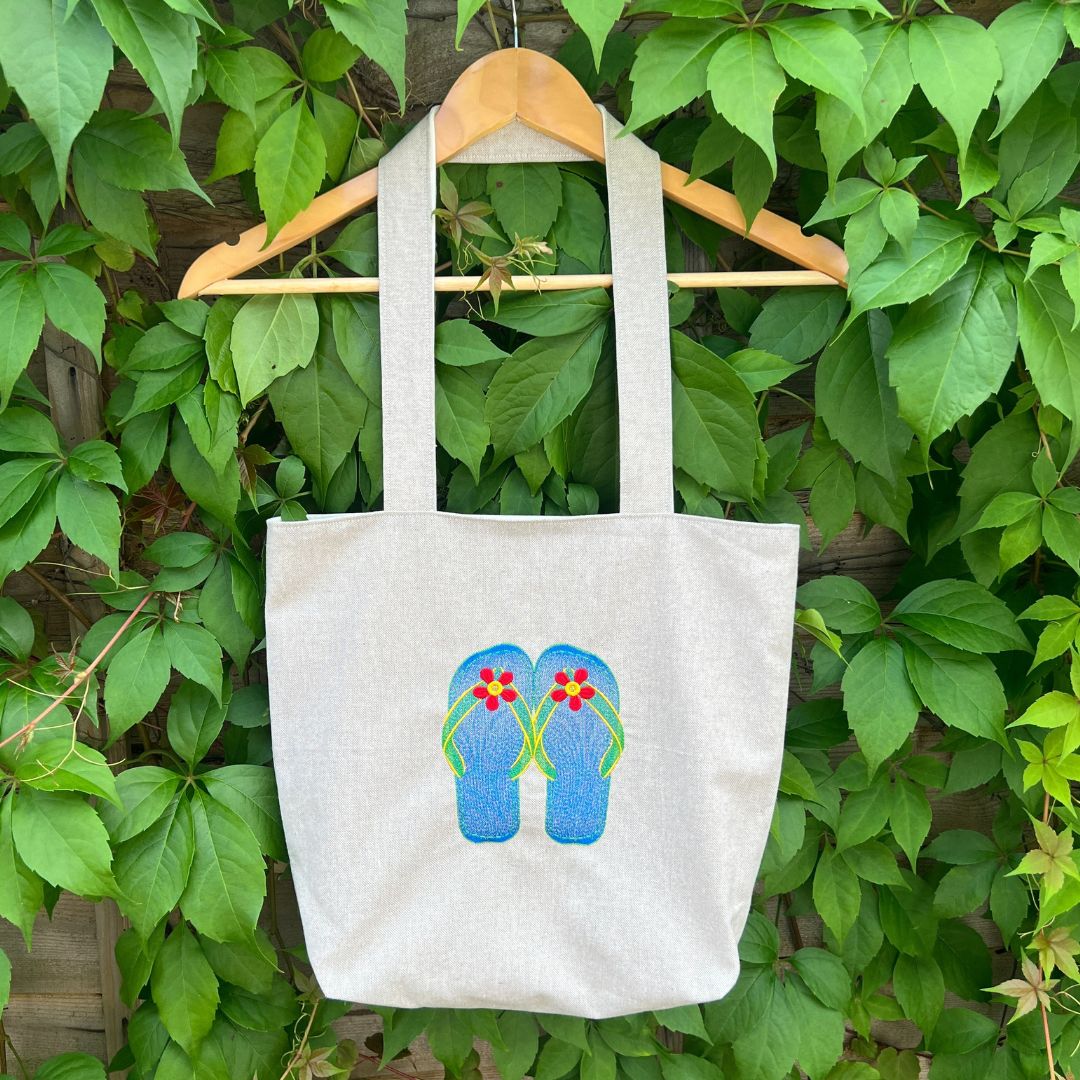Tote bag with flip-flop design hanging on a hanger against a green leafy background