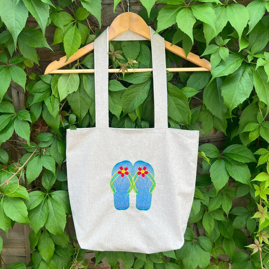 Tote bag with flip-flop design hanging on a hanger against a green leafy background