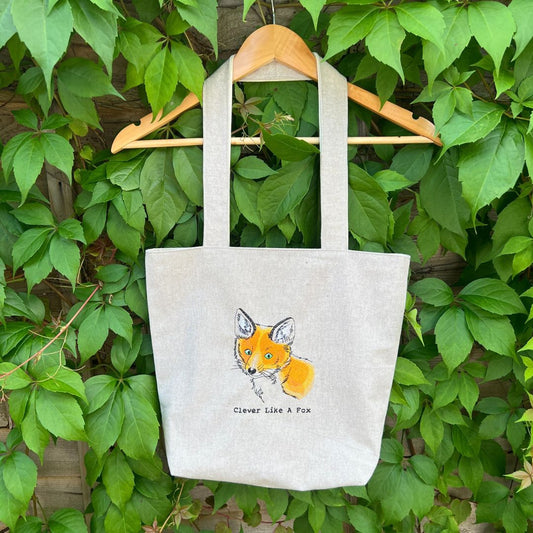 Tote bag with fox design and text 'Clever Like A Fox' hanging on a wooden hanger against a green leafy background.