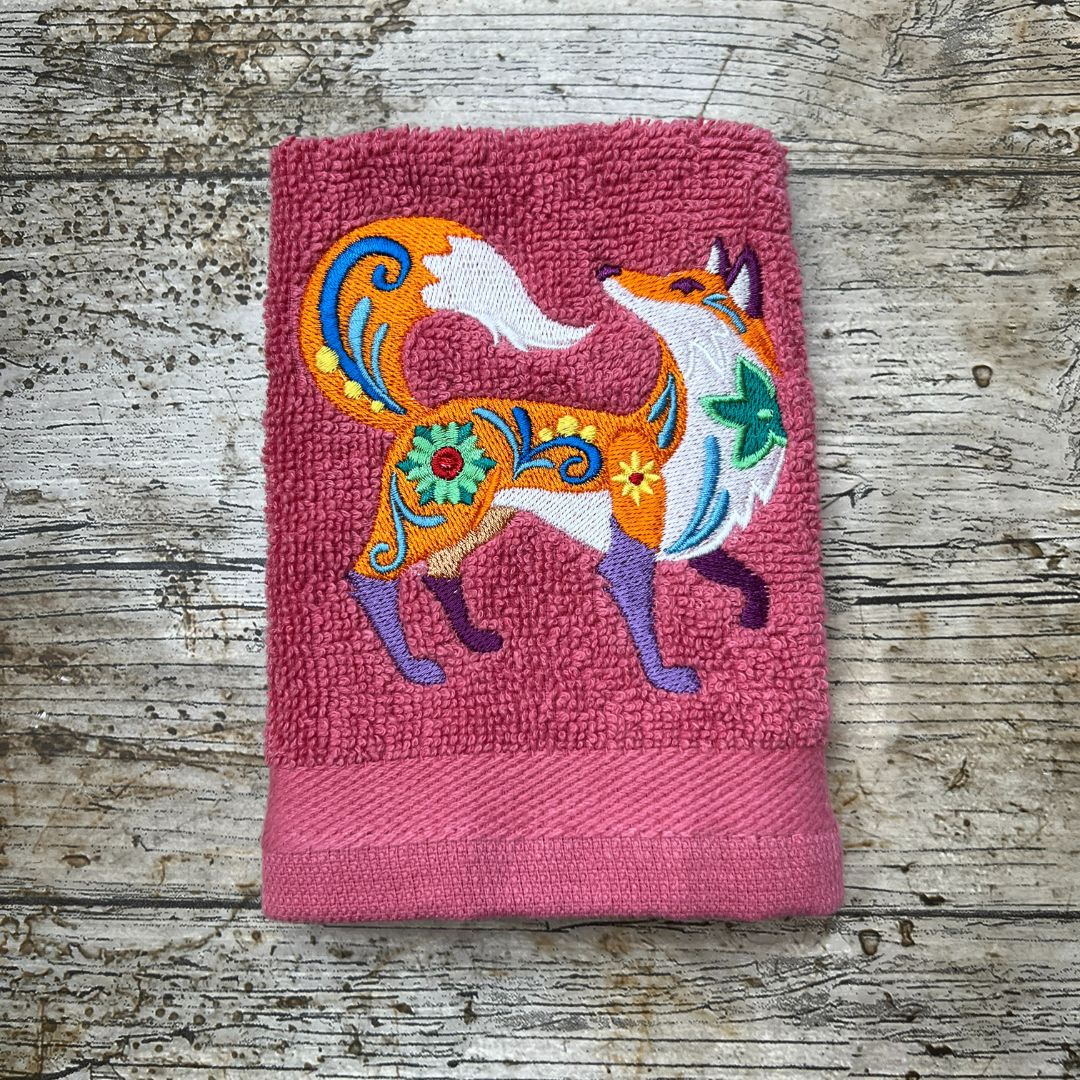 a Funky fox embroidered onto a  face cloth and place on a wooden background, made by Coralie's crafts & Gifts in Dorset.