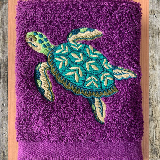 A purple terry cloth towel featuring an embroidered sea turtle design in teal, green, and cream, displayed flat on a wooden surface.