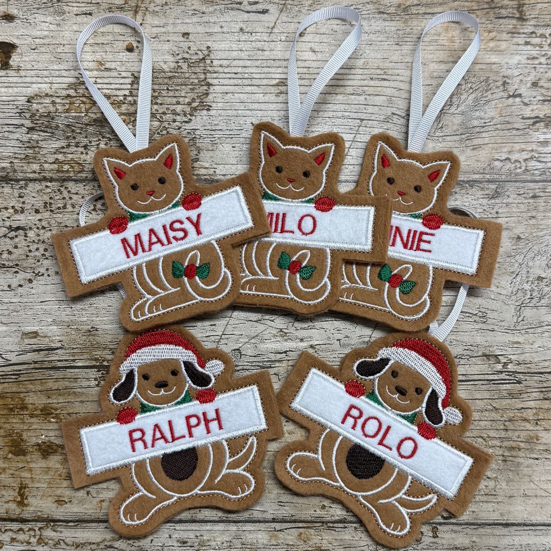 Six personalized Christmas embroidered Gingerbread ornaments shaped like a cat and a dog, arranged on a rustic wooden surface. The ornaments are brown with white hanging ribbons, featuring custom names (MAISY, MILO, NINE, RALPH, ROLO) in red embroidery on white banners, along with small red and green accents.