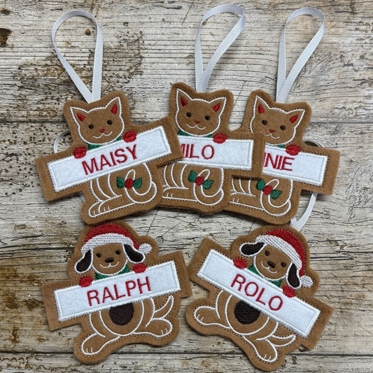 Six personalized Christmas embroidered Gingerbread ornaments shaped like a cat and a dog, arranged on a rustic wooden surface. The ornaments are brown with white hanging ribbons, featuring custom names (MAISY, MILO, NINE, RALPH, ROLO) in red embroidery on white banners, along with small red and green accents.