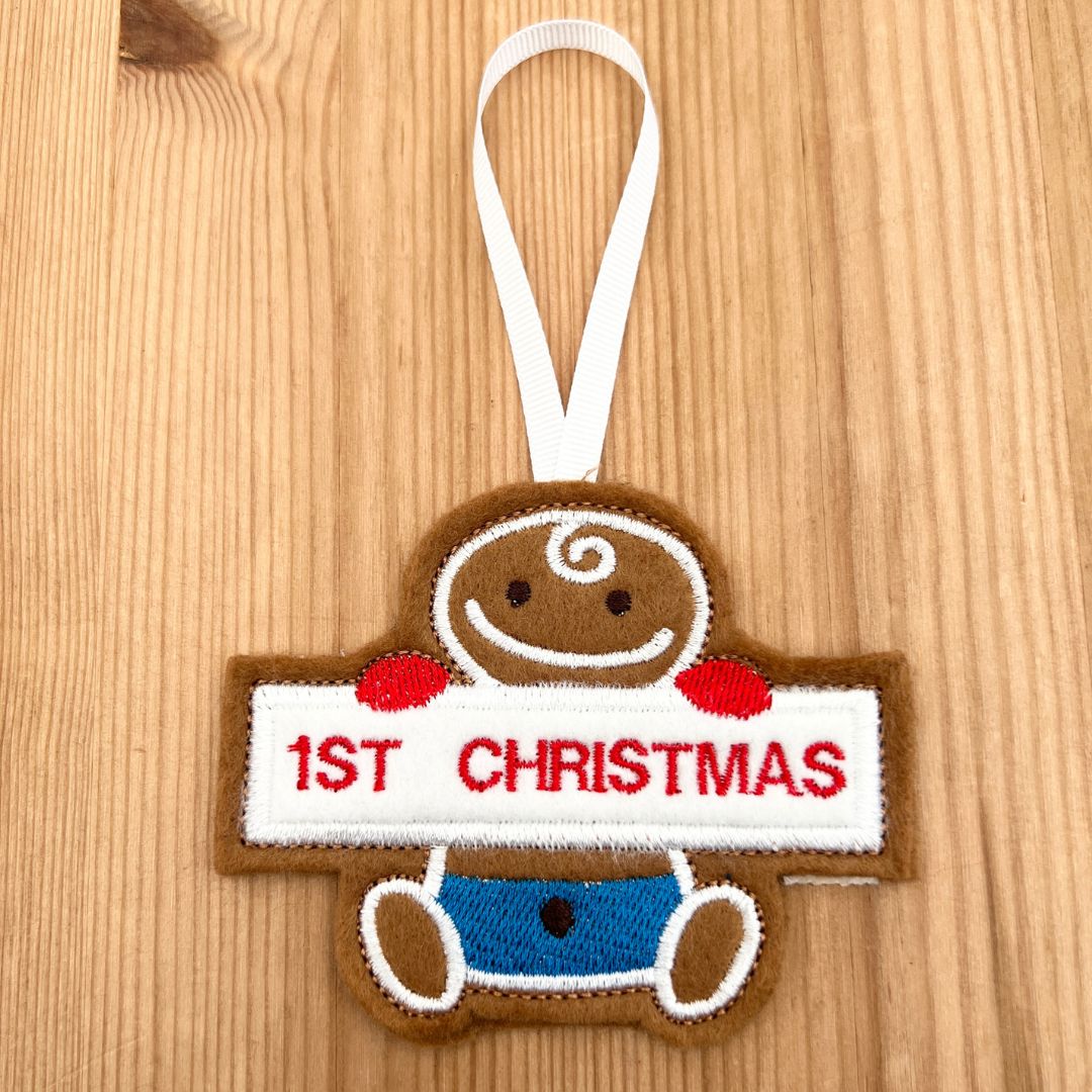 A handmade felt Christmas gingerbread ornament shaped like a gingerbread baby holding a white banner that reads '1ST CHRISTMAS' in red embroidery, with a white hanging loop, shown on a light wood background.