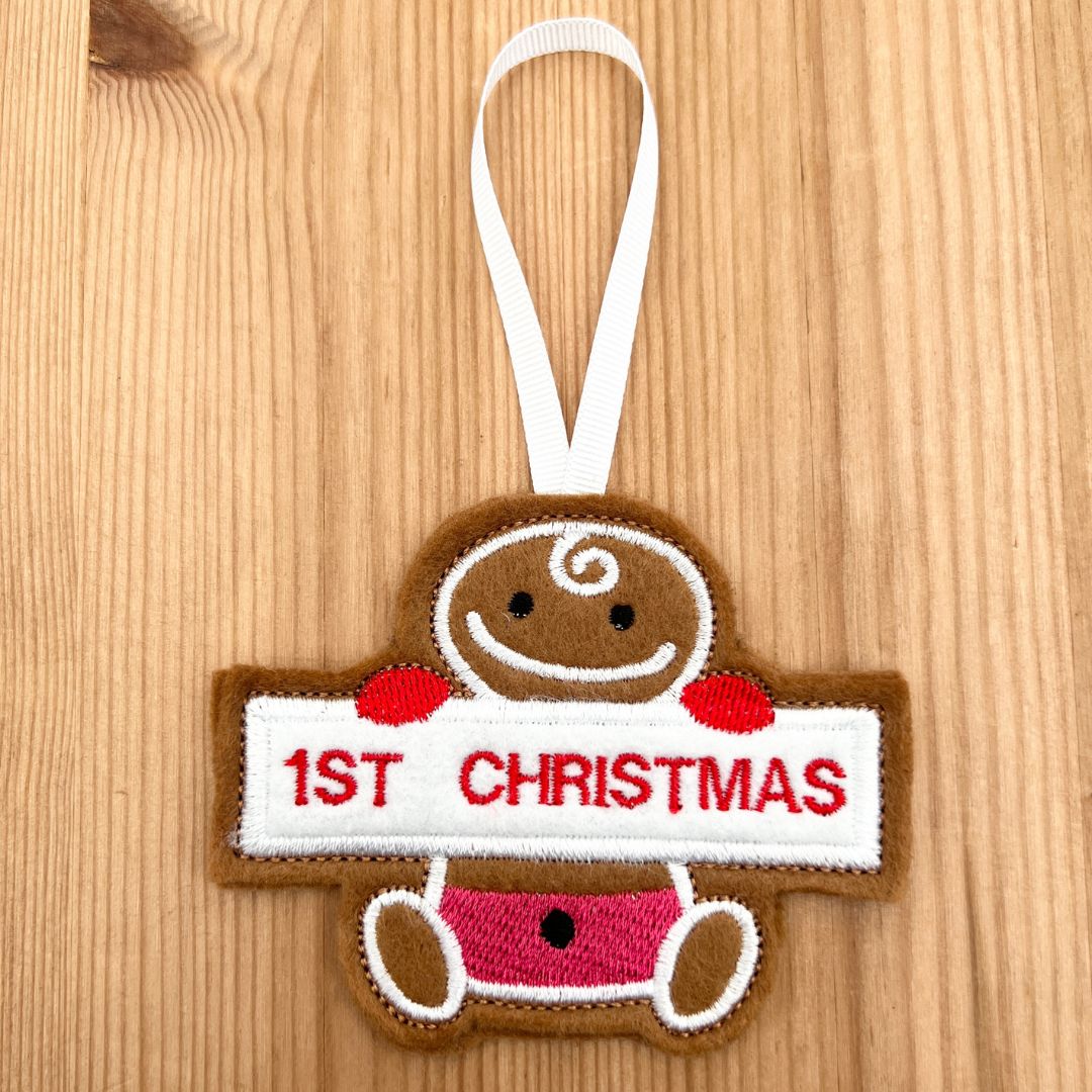 A gingerbread man felt Christmas  gingerbread baby ornament hanging on a light wood background. The ornament is brown with white, red, and pink details, holding a white banner that reads '1st Christmas' in red letters, and has a white hanging loop at the top.