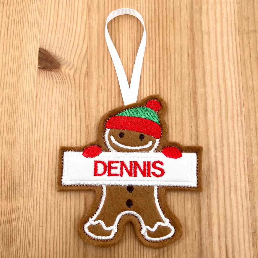 A personalized gingerbread boy Christmas ornament on a light wood background. The ornament is brown with a white hanging ribbon, featuring a green and red striped hat, red mittens, and a white banner with the name 'DENNIS' in red embroidered letters.