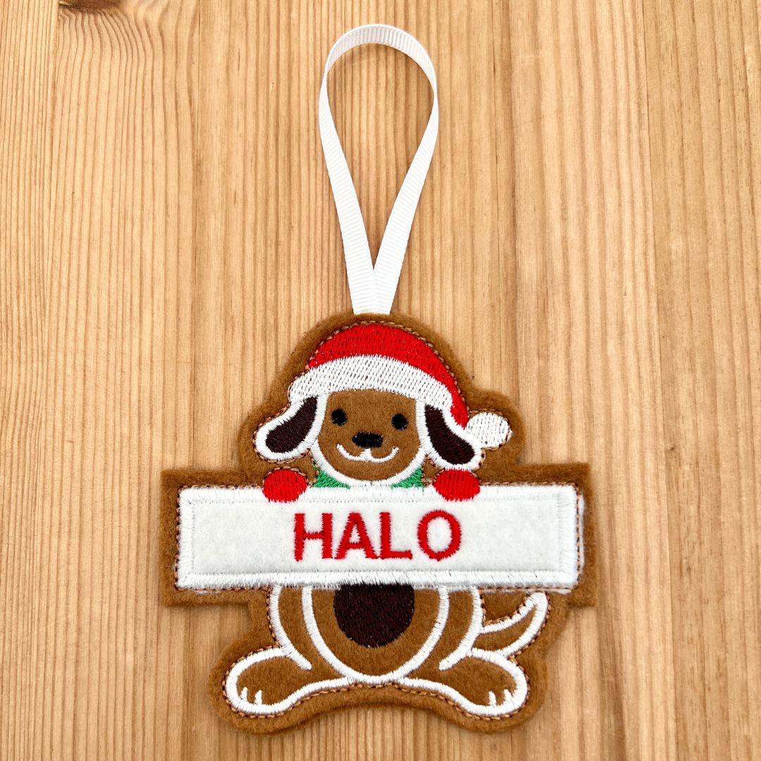 A felt Christmas gingerbread ornament shaped like a dog wearing a Santa hat and holding a white banner that says 'HALO' in red letters, hanging from a white ribbon on a light wood background.