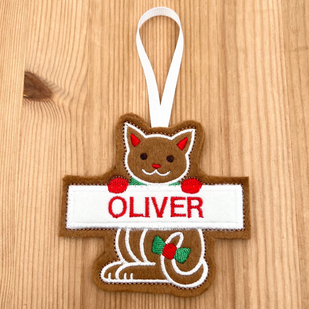 A personalized felt Christmas ornament shaped like a gingerbread cat holding a white sign with the name 'OLIVER' in red embroidery, hanging on a light wood background.